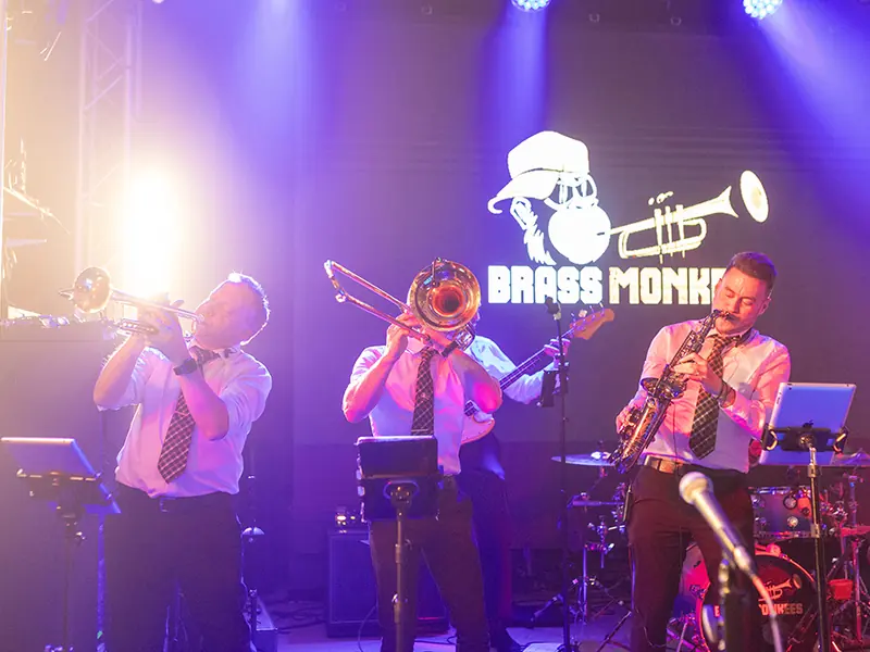 Brass Monkees horn section playing trumpet, trombone and sax in front of the band logo
