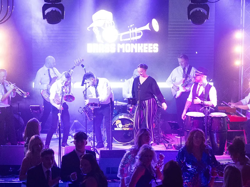 The full Brass Monkees 9-piece band on stage with horns, singers and percussion in front of the band logo